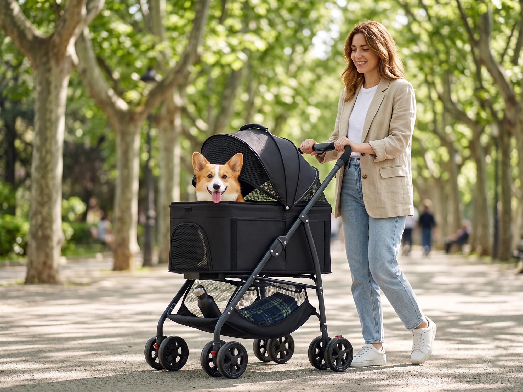 A smiling young woman walks down a sunny, tree-lined park path pushing a black multi-function pet stroller. A happy Corgi dog sits inside the stroller's open mesh carrier, and the bottom storage basket holds a water bottle and a folded plaid blanket.