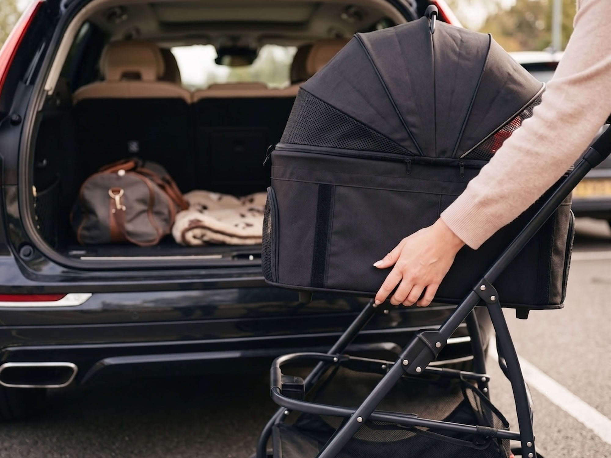 A person's hand lifting a black, detachable pet carrier off its stroller frame. In the background, a dark SUV has its trunk open, revealing a brown duffel bag and a patterned blanket inside, suggesting they are packing for a trip.