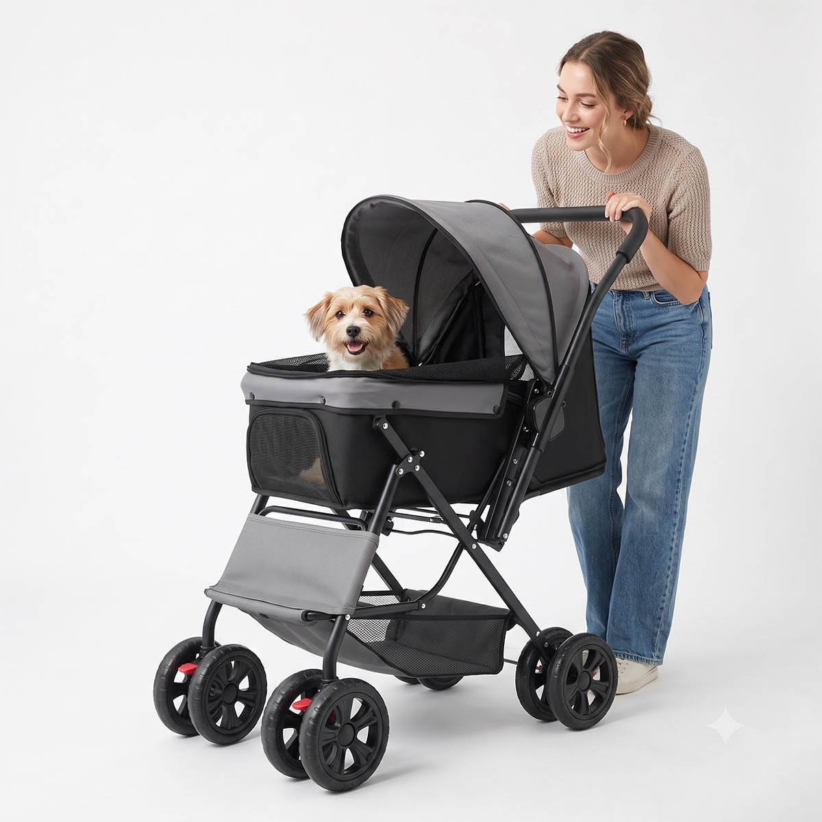 A young European-American woman pushing a gray and black pet stroller against a plain white background. Inside the open-canopy stroller basket, a happy small brown and white dog sits looking forward. The stroller features a black frame, mesh side panels, double front wheels, and a large bottom storage basket.